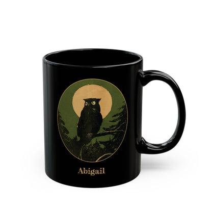A black ceramic 11oz mug featuring a circular graphic of a black owl perched on a branch in front of a tan moon, with the name 'Abigail' printed below in a gold-like color, on a white background. Right handle view