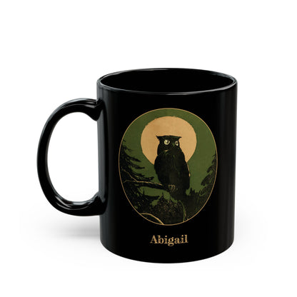 A black ceramic mug featuring a circular vintage-style illustration of an owl perched on a branch against a green background with a tan moon, and the name 'Abigail' printed below in a gold-like color.