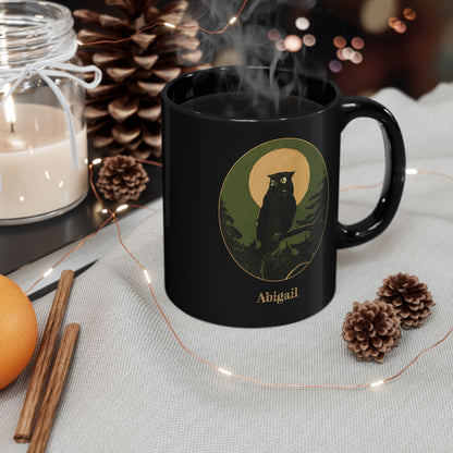 A black ceramic 11oz mug with a handle, featuring a graphic of an owl in a tree silhouette against a gold color and green circular background with the name 'Abigail' printed below. The mug is styled on a light fabric surface with a candle, pinecones, cinnamon sticks, and fairy lights.