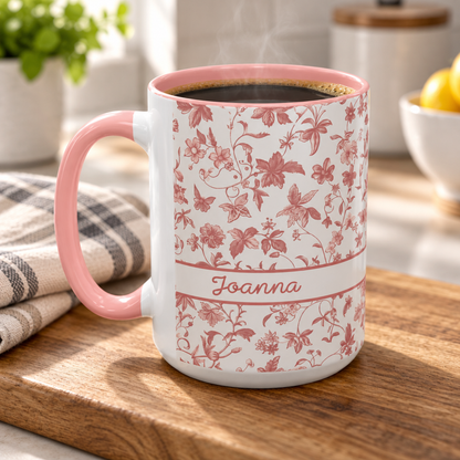 A white ceramic 15oz coffee mug with a pink floral chinoiserie pattern and the name 'Joanna' printed on the front, filled with coffee and set on a wooden cutting board in a kitchen setting.