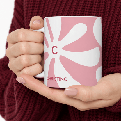A close-up of a person holding a white ceramic mug with a pink and white abstract floral pattern, featuring the initial 'C' and the name 'CHRISTINE' printed on it. The person is wearing a dark red knit sweater.