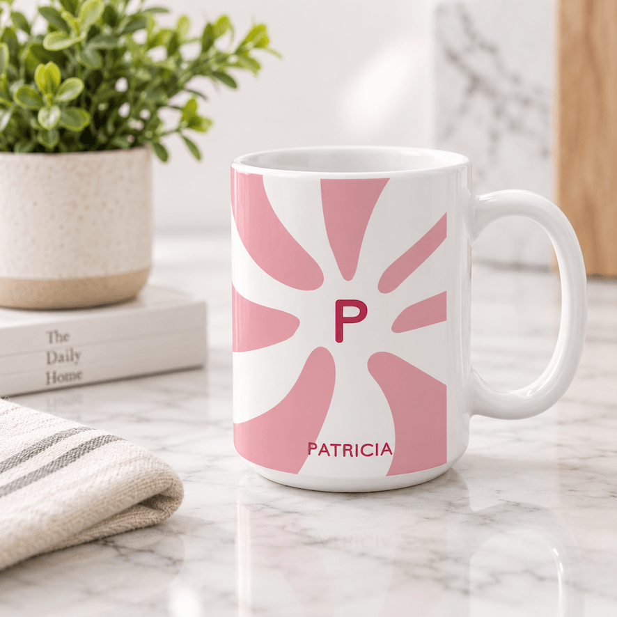 A white ceramic mug with a pink sunburst pattern featuring the initial 'P' and the name 'PATRICIA' printed in pink, set on a marble countertop with a potted plant and books in the background.