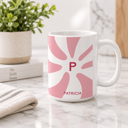 A white ceramic mug with a pink sunburst pattern featuring the initial 'P' and the name 'PATRICIA' printed in pink, set on a marble countertop with a potted plant and books in the background.