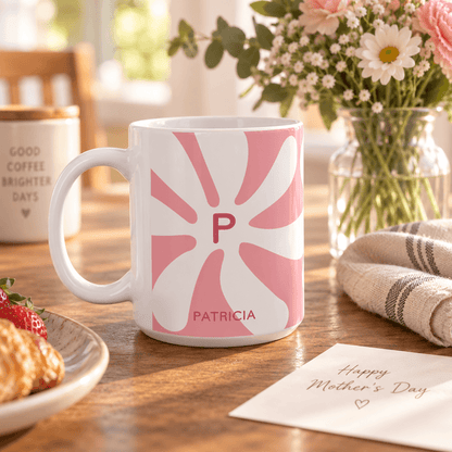 A white ceramic mug with a pink sunburst pattern featuring the initial 'P' and the name 'PATRICIA' printed in pink, styled on a wooden table with a Mother's Day card, flowers, and a cozy kitchen background.