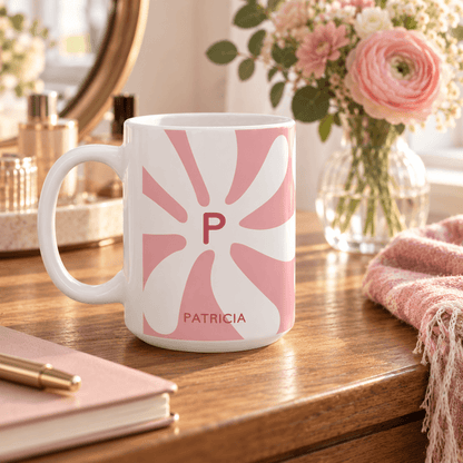 A white ceramic mug with a pink retro sunburst pattern featuring the initial 'P' and the name 'PATRICIA' printed in pink, styled on a wooden desk with a notebook, pen, and a vase of pink flowers in the background.