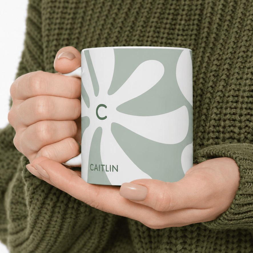 A person in an olive green knit sweater holds a white ceramic 11oz mug with a sage green and white retro flower pattern, featuring a green letter 'C' and the name 'CAITLIN'.