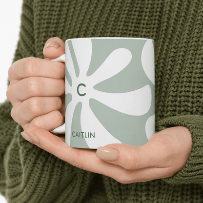 A person in an olive green knit sweater holds a white ceramic 11oz mug with a sage green and white retro flower pattern, featuring a green letter 'C' and the name 'CAITLIN'.
