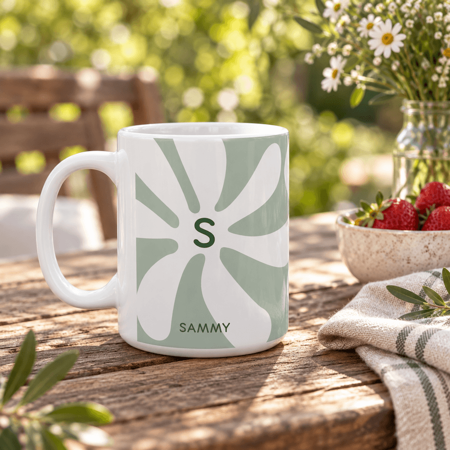 A white ceramic 15oz mug with a sage green retro sunburst pattern featuring the initial 'S' and the name 'SAMMY' printed at the bottom, set on a rustic wooden table in an outdoor garden setting.