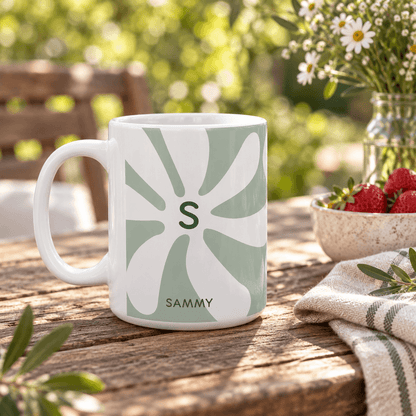 A white ceramic 15oz mug with a sage green retro sunburst pattern featuring the initial 'S' and the name 'SAMMY' printed at the bottom, set on a rustic wooden table in an outdoor garden setting.