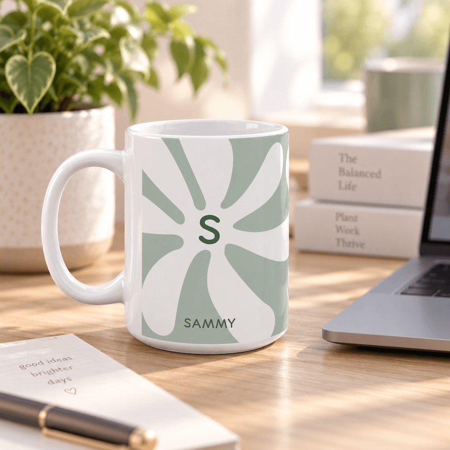 A white ceramic 15oz mug with a sage green and white sunburst pattern, featuring a green monogram letter 'S' and the name 'SAMMY' printed at the bottom. The mug is set on a wooden desk next to a laptop, notebook, and potted plant.