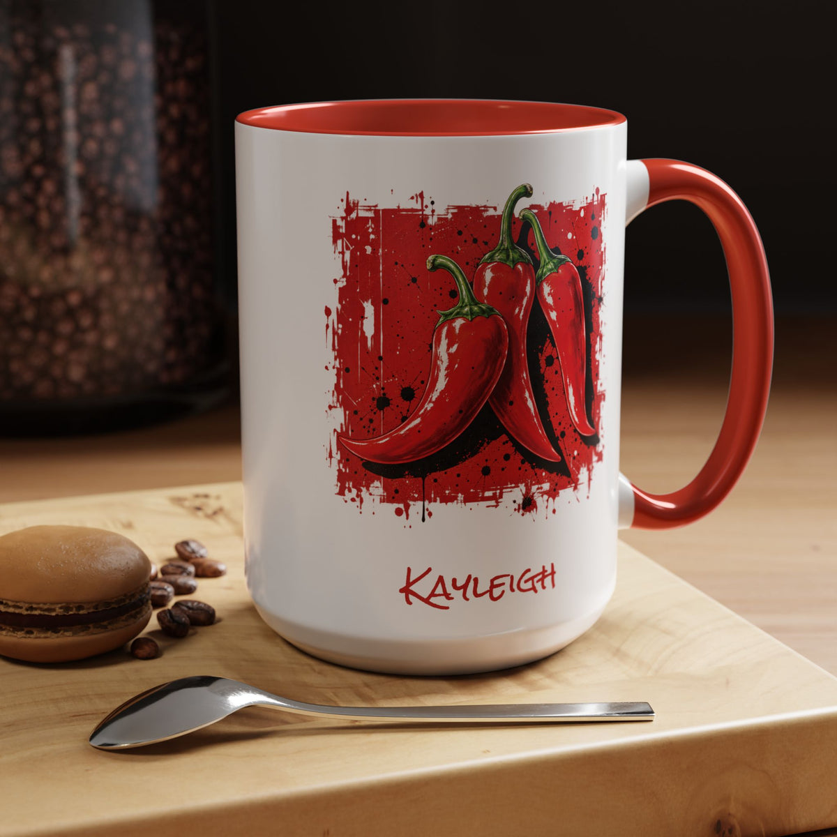 A white ceramic mug with a red interior, red handle, and a red graphic of 3 chili peppers, with the name 'KAYLEIGH' printed in red near the bottom. The mug sits on a wooden board with a spoon and a macaron in the foreground, with a dark background.