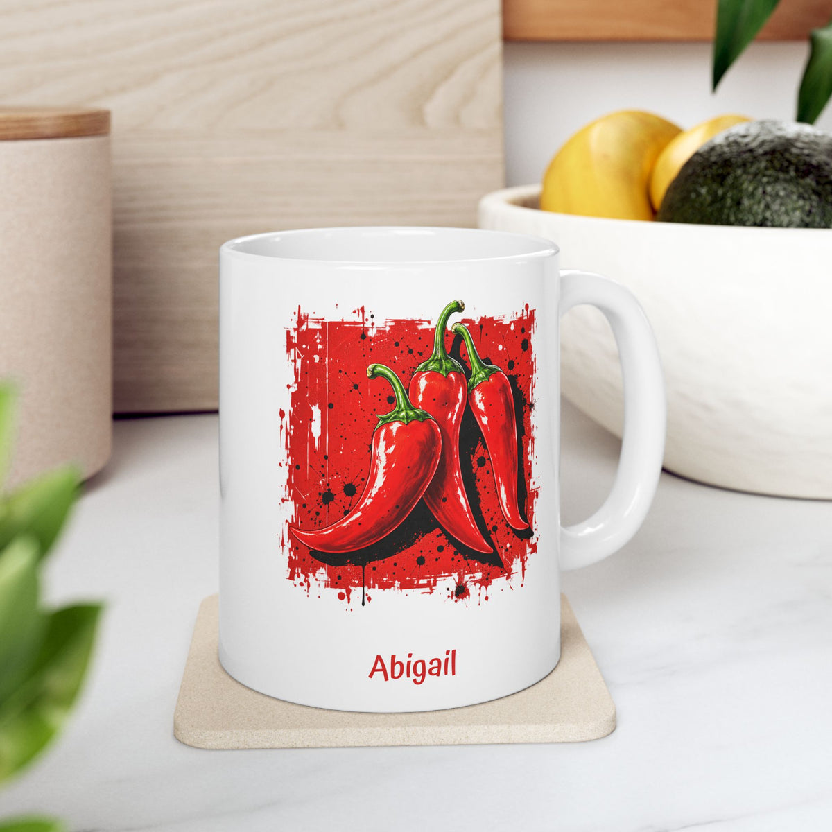 A white ceramic 11oz mug with a graphic of three red chili peppers on a red paint-splatter background, with the name 'Abigail' printed in red below. The mug is sitting on a light beige coaster on a white marble countertop with a wooden wall and a bowl of fruit in the background.