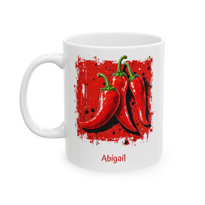 A white ceramic 11oz mug featuring a graphic print of three red chili peppers on a red splattered background, with the name 'Abigail' printed in red below. Left handle view.