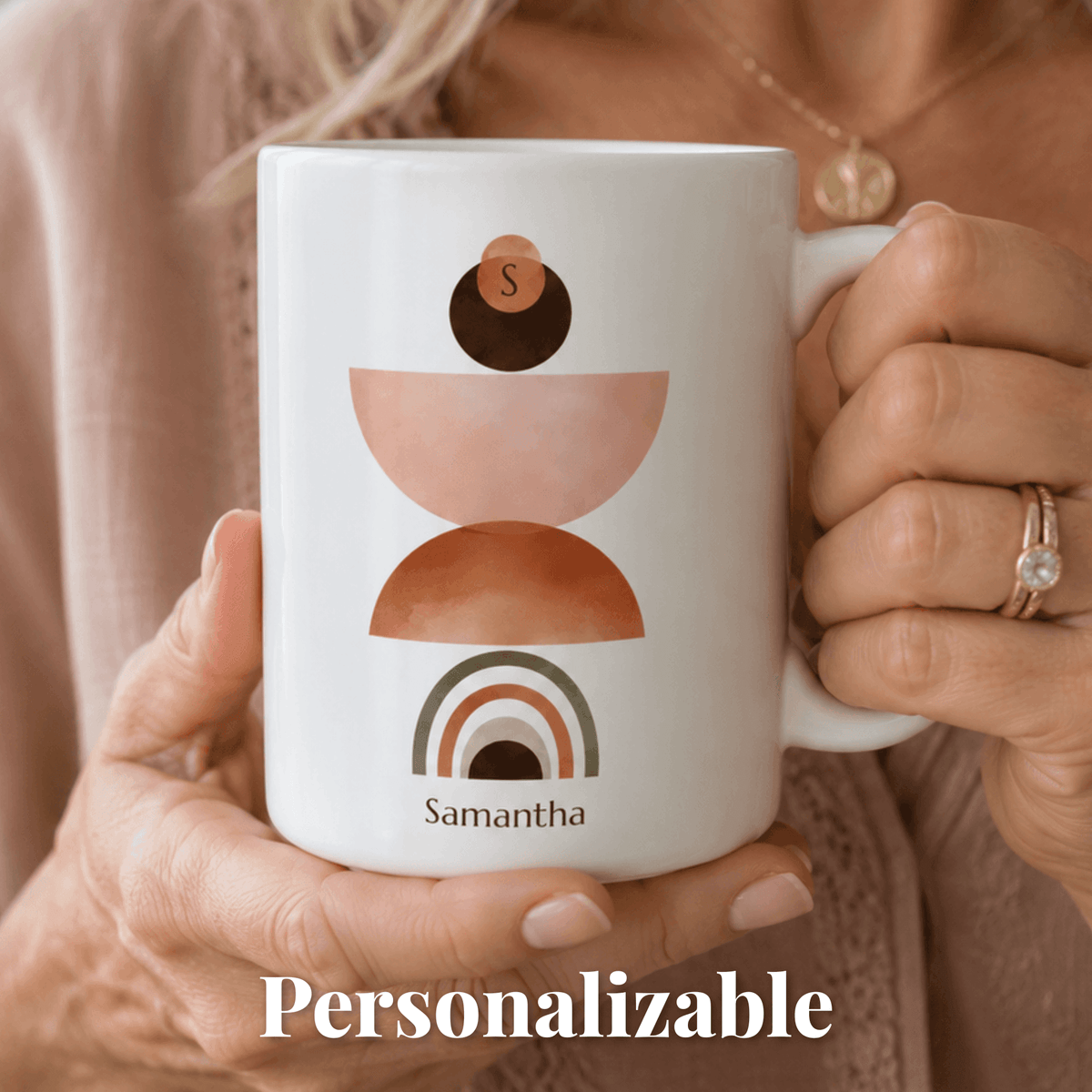 A person holds a white ceramic mug with a minimalist stacked-geometric design in blush pink, terracotta, and dark brown, plus the personalized name 'Samantha' and the word 'Personalizable' overlaid at the bottom.