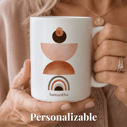 A person holds a white ceramic mug with a minimalist stacked-geometric design in blush pink, terracotta, and dark brown, plus the personalized name 'Samantha' and the word 'Personalizable' overlaid at the bottom.