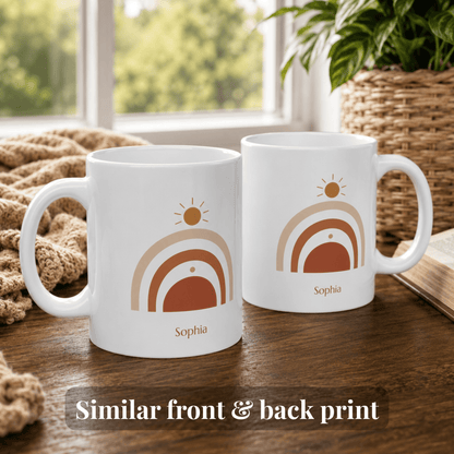 Two white 11oz ceramic mugs with a matching minimalist sun and rainbow design, labeled 'Sophia' on the front, displayed on a wooden table in a cozy room setting with a window, plant, and blanket in the background.