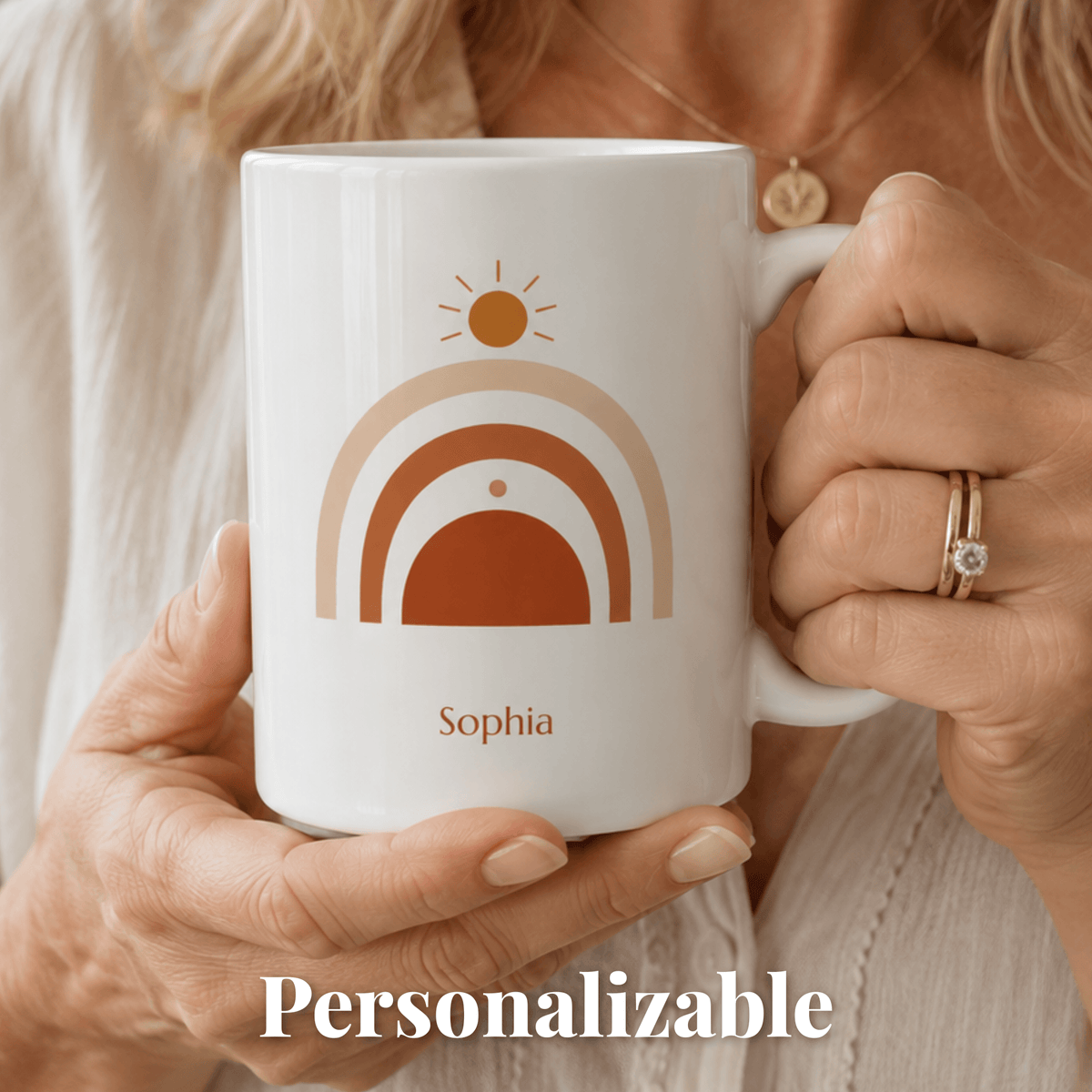 A person holds a white ceramic mug with a retro-style rainbow design in beige, tan, and rust orange, with the name 'Sophia' printed below. The mug is shown close-up with the word 'Personalizable' overlaid at the bottom.