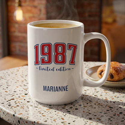 A white ceramic coffee 15oz mug with '1987 limited edition' and the name 'MARIANNE' printed on it, sitting on a speckled countertop with a croissant in the background. Right handle view.
