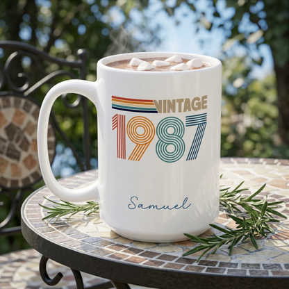 A white ceramic 15oz mug with a retro 1987 design and the name 'Samuel' printed on it, filled with hot chocolate and marshmallows, set on a mosaic table outdoors. Left handle view.