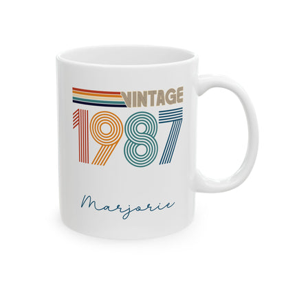 A white ceramic 11oz mug featuring a retro-style 'VINTAGE 1987' graphic with colorful stripes and the name 'Marjorie' written in cursive below, shown on a clean white background. Right handle view.