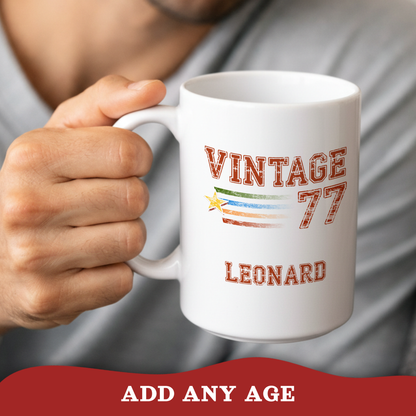 A person holds a white ceramic mug with a custom printed design that reads 'VINTAGE 77 LEONARD' with a retro flag motif. The background shows a blurred gray shirt and skin, with a red banner at the bottom reading 'ADD ANY AGE'.