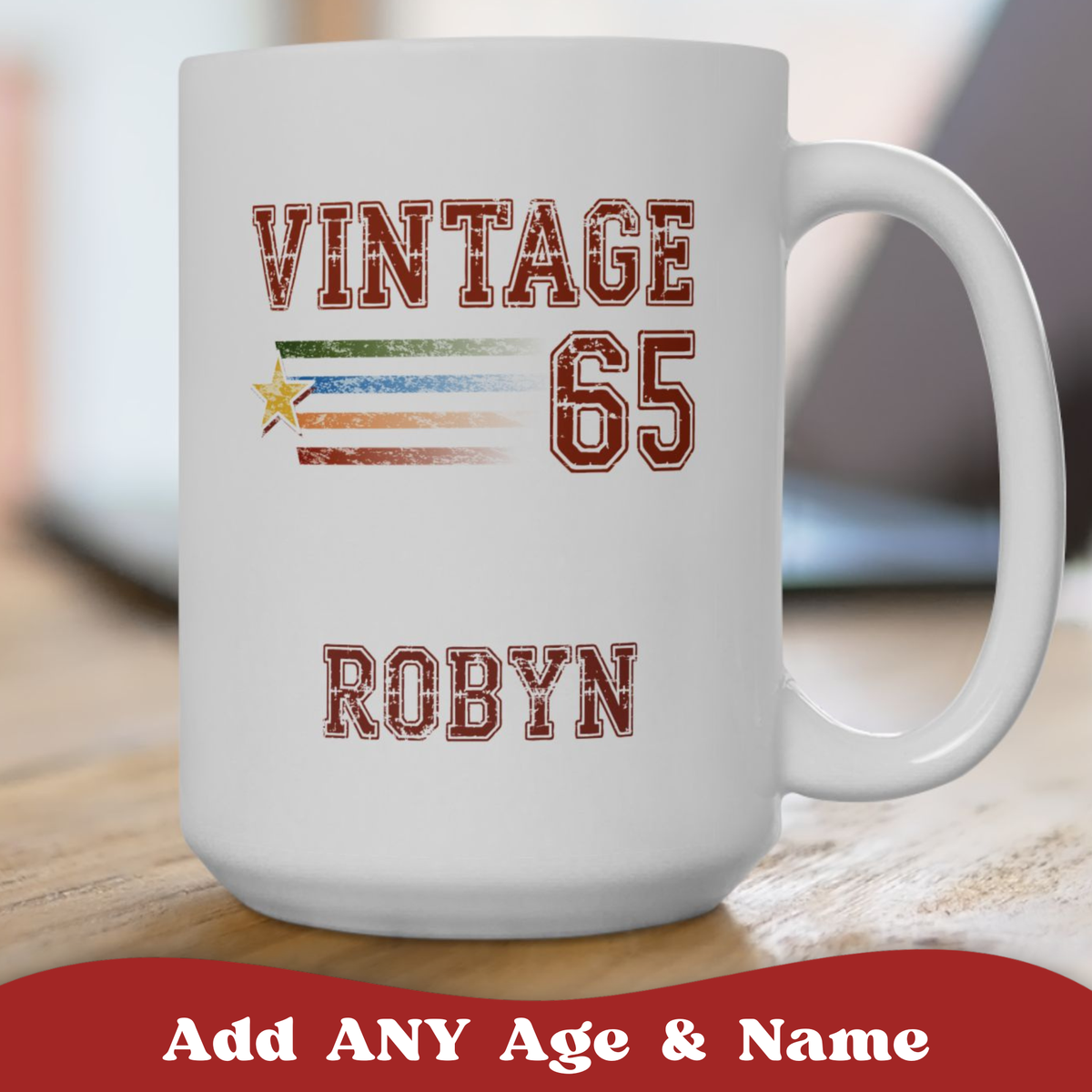 A white ceramic 15oz mug with a custom sports-style graphic reading 'VINTAGE 65 ROBYN' in distressed red lettering, featuring a horizontal multicolor stripe and star motif. The mug sits on a wooden surface with a blurred indoor background.