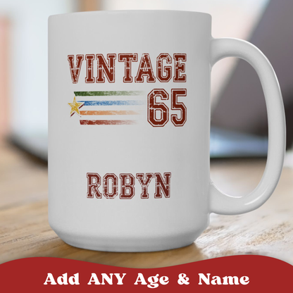 A white ceramic 15oz mug with a custom sports-style graphic reading 'VINTAGE 65 ROBYN' in distressed red lettering, featuring a horizontal multicolor stripe and star motif. The mug sits on a wooden surface with a blurred indoor background.