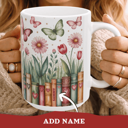 A white ceramic 15oz mug held by a person, featuring a printed design of vintage book spines decorated with hearts, with flowers and butterflies growing out of them, on a white background. A subtle personalizable name on one of the book spines.