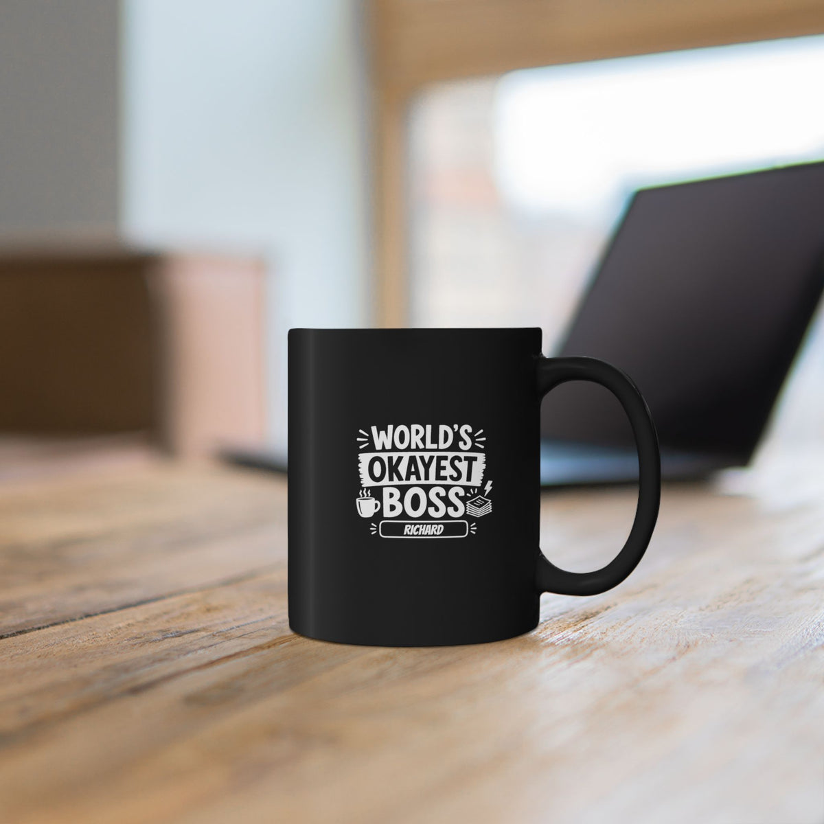 Black 11oz mug with white illustrative typographic design saying 'World's Okayest Boss' and a personalized name at the bottom, left handle view. Mug is placed on a wooden table with blurred laptop in the background.