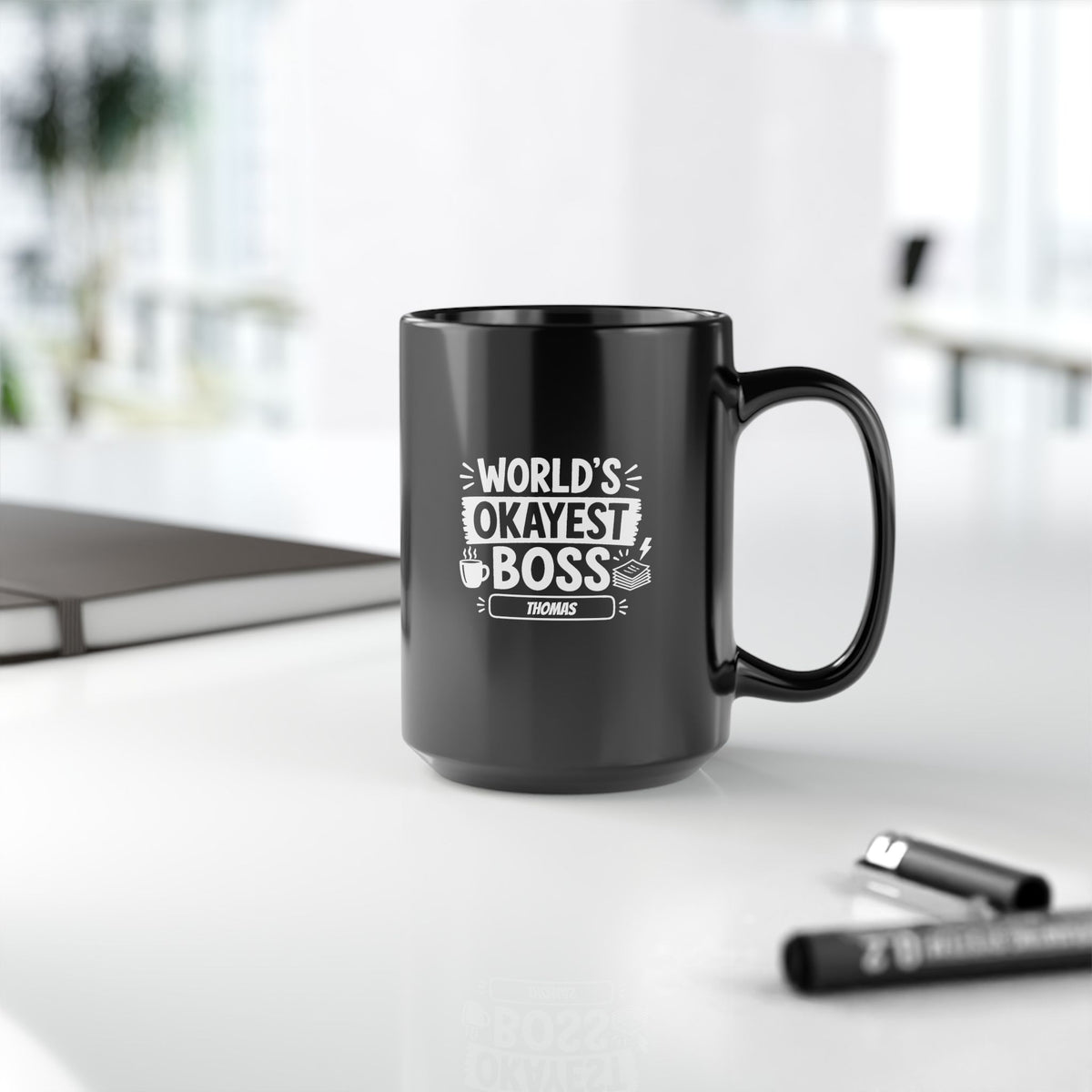 Glossy black 15oz mug with white illustrative typographic design saying 'World's Okayest Boss' and a personalized name at the bottom, right handle view. Mug is placed on a white surface with blurred pen and a notebook.