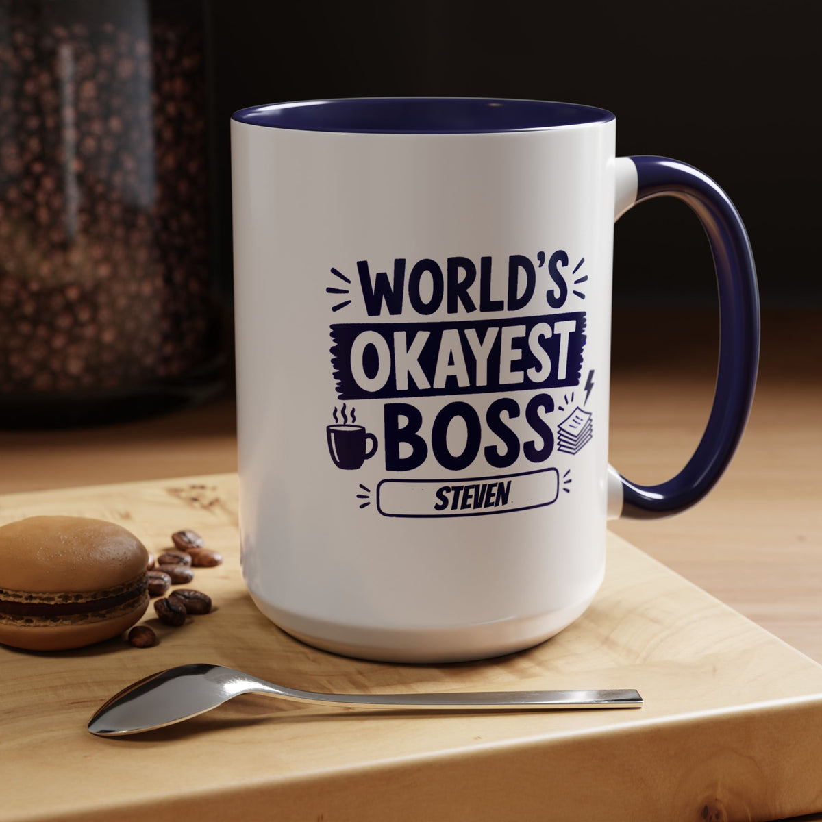 A white ceramic 15oz coffee mug with a navy blue handle and interior, featuring the text 'WORLD'S OKAYEST BOSS STEVEN' in navy blue. The mug is placed on a wooden cutting board with a spoon, coffee beans, and a macaron in a warm, cozy setting.