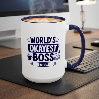 A white ceramic 15oz coffee mug with a navy handle and rim, printed with the text 'WORLD'S OKAYEST BOSS STEVEN' in navy. The mug is filled with coffee and sits on a black desk mat on a wooden desk next to a keyboard and computer monitor.